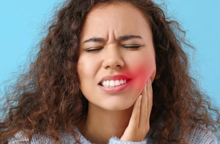Woman with curly hair wincing with her eyes shut, pressing a hand to her cheek, with the jaw area highlighted in red to show toothache